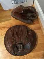 Photo of two vintage leather baseball mitts on wooden floor showing full mitts and details of leather wear and lacing.