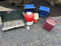 Overview of the Coleman camping equipment lot on asphalt ground with mulch background. Includes stove, chairs, beverage jug, lantern, and propane tank case.
