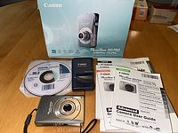 Canon PowerShot SD750 camera with battery charger, Solution Disk, instruction booklets, and original box visible on wooden surface.