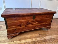 Closed cedar chest showing natural wood grain, metal corner brackets, and decorative feet with wheels beneath.