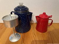 Blue speckled enamelware percolator coffee pot and red speckled enamelware coffee pot with lids, displayed with metal coffee basket insert.