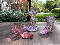 Three pieces: bridesmaid figurine, glass boot, and glass shoe laid out on a table outside with greenery background