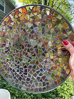 Front view of round glass mosaic garden plate with orange and amber tones held up outdoors.