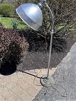 Full view of the industrial adjustable height floor lamp positioned outdoors on pavement and near shrubbery.