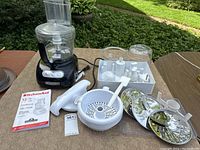 Food processor base unit with multiple accessories including discs, straining bowl, spatula, tamper tool, and instruction manual, all shown arranged on a flat surface outdoors.