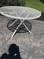 Outdoor image of a white metal circular patio table with mesh top and crossed tubular legs, central hole for umbrella visible.