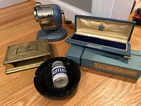 Overview photo showing all items, including the Goodyear note holder, Boston pencil sharpener, glass Bufferin bottle inside black ashtray, and Hamilton watch box with packaging.