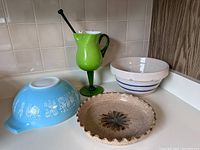 Four items displayed together on counter: Pyrex bowl face-down showing pattern, green glass pitcher, Schucharatt pie plate front, farmhouse bowl