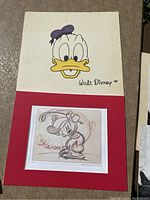Photo showing both Donald Duck colored signed drawing and Mickey Mouse pencil sketch mounted on red mat
