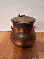 Front view of aged copper pot with lid. Pot shows two side handles and stamped markings visible on the front.