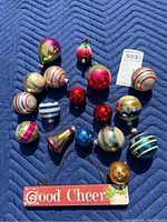 Top-down view of the collection of vintage Christmas ornaments arranged on a blue textured blanket showing a variety of colors and shapes including stripes, speckles, and solid colors, along with a "Good Cheer" holiday sign.