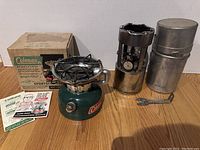 Overview of two Coleman single burner stoves, one WWII silver model with funnel, one green Sportster model with original box and manuals, placed on wood floor.