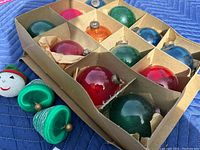Photo showing original box with divided compartments containing multiple spherical glass Christmas ornaments in green, red, blue, pink, and orange. Includes a small white snowman face ornament and two green felt Christmas tree shaped ornaments.