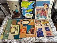 Collection of vintage advertisement artwork and posters displayed over a table including travel, cosmetics, photo processing, and footwear ads.