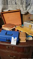 Photo showing an assortment of art boxes and supplies arranged on a wooden surface beneath a white lace curtain.