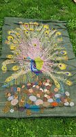 Full view of embroidered peacock on burlap fabric lying on grass, showing vibrant colors and detailed stitching across entire textile.