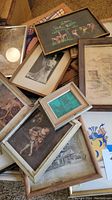 Top view of assorted framed artworks piled, showing various artworks including a colorful animal scene on black background and sepia drawings