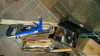 Overview of two cardboard boxes and one metal toolbox all containing assorted tools including saw, caulking gun, and hand tools