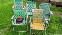 Set of five midcentury basket weave folding outdoor chairs arranged on lawn, showing woven plastic seats and backs in green, yellow, beige, and blue, with aluminum frames and extra strapping rolls on one chair.