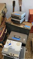 Photo showing two acoustic guitars, vintage music players, reel-to-reel tape recorder, amplifier and equipment cases arranged together.