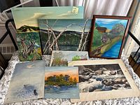 Overview of all seven nature themed paintings: various sizes and some framed, laid out on a white patterned sheet. Scenes include mountains, trees, water, buildings, and rocks.