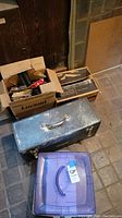 Photo of large black metal toolbox, cardboard boxes, and purple lid plastic storage container on the ground.