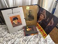 Photo showing the large spiral-bound Van Gogh book, two large prints/posters, and a hardcover Van Gogh art book arranged on a bed.
