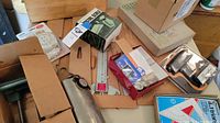 Overview of several woodworking and art supplies in cardboard boxes including clamps, rulers, and packaged accessories