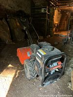 Orange yard tiller with visible Subaru branding on the engine, black protective cage around the engine and big wheels, photographed from side and front angles in a dim basement environment.