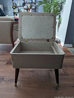 Top-front view of mid century footstool open showing storage compartment inside and tapered wooden legs with brass tips.