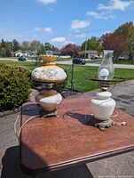 Two antique lamps on a table outside: the porcelain Gone with the Wind lamp is on the left with painted floral decoration and beige top; the white milk glass swirl lamp with glass chimney is on the right.
