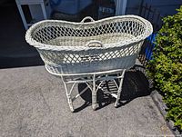 Photo showing oval light-colored wicker bassinet with lattice weave and steel rod stand with wheels, placed outside near a building.