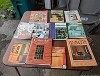 Eleven books displayed on a table outdoors focused on home design, carpentry, landscaping, old houses, and decorating.