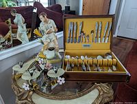 Photo showing Erza Brooks decanter with female figurine and missing tennis racket piece, vintage Community silverware set in wood box, and Mid Century toleware flower sconce with some flowers and leaves.