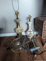 Two Mid Century handpainted porcelain lamps featuring gold gilt and floral designs alongside one heavy leaded blown glass hanging lamp on wooden floor.