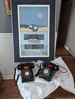 Photo shows framed mixed media artwork by Eric Bellman alongside two black vintage rotary dial telephones, placed on a white fabric.