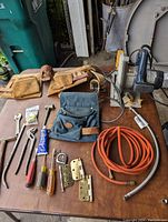 Full lot showing green Ryobi saw, assorted hand tools, metal hinges, two toolbelts, and orange coiled hose on wooden table.