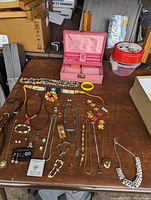 Photo of pink vintage musical jewelry box and various costume jewelry including bolo ties, necklaces, bracelets, pins, and earrings laid out on a table.