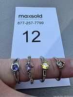 Four rings shown on fingers with a MaxSold lot card behind, highlighting the colored stones and rhinestones on the rings.