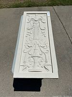 Photo of one rectangular decorative wall panel with intricate relief floral and ornamental design, cream or off-white, placed on outdoor table.