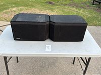 Pair of black Bose 301 Series II speakers positioned side by side on white folding table outdoors, front view with cloth covers visible.