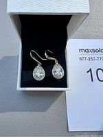 Pair of gold-tone earrings with large teardrop-shaped clear stones and smaller rhinestones, displayed in small white box with black interior.