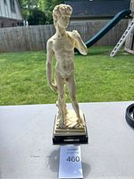 Full frontal photo of Michelangelo's David replica sculpture on black base displayed outdoors on white surface with grass and playground background.