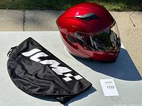 Red ILM helmet with black bag and auction lot card on table outdoors under daylight