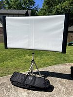 Front view of the portable screen fully mounted on tripod stand with carrying bag below on concrete surface outdoors.