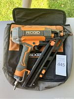 Ridgid nailer placed on black carrying bag, showing the orange and gray body and front metal magazine.