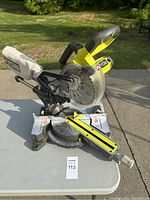 Front view of the Ryobi compound miter saw on a table outdoors with dust collection bag attached.