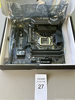 Motherboard shown in box with protective black plastic inside a cardboard box with maxsold lot tag.