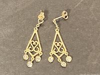 Pair of gold dangle earrings shown on gray background displaying front view and side angle with openwork heart pattern and three clear bezel-set stones hanging below