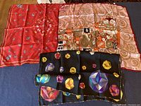 Three scarves laid flat showing red floral scarf, beige with abstract artwork, and black with geometric shapes